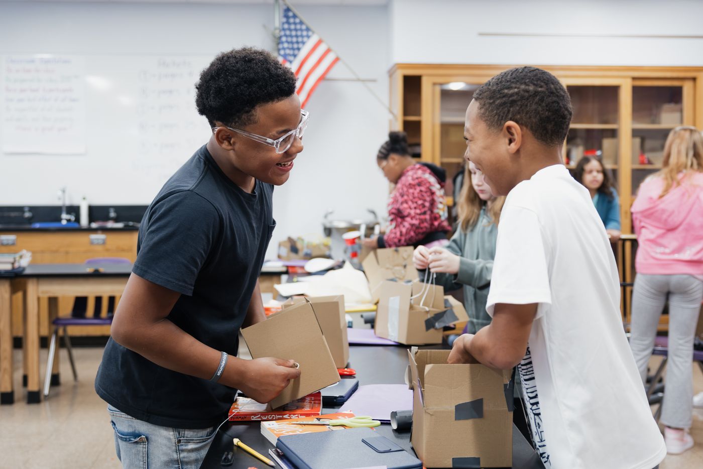 Two students grin at each other while assembling cardboard boxes in a science lab.