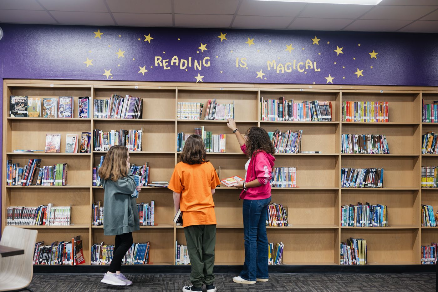 Three students browse the shelves beneath a mural reading Reading Is Magical.