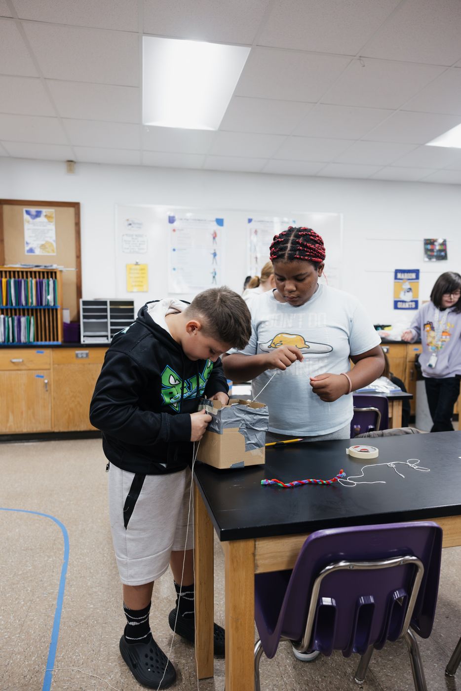 Two students work together on a taped cardboard build at a lab table.