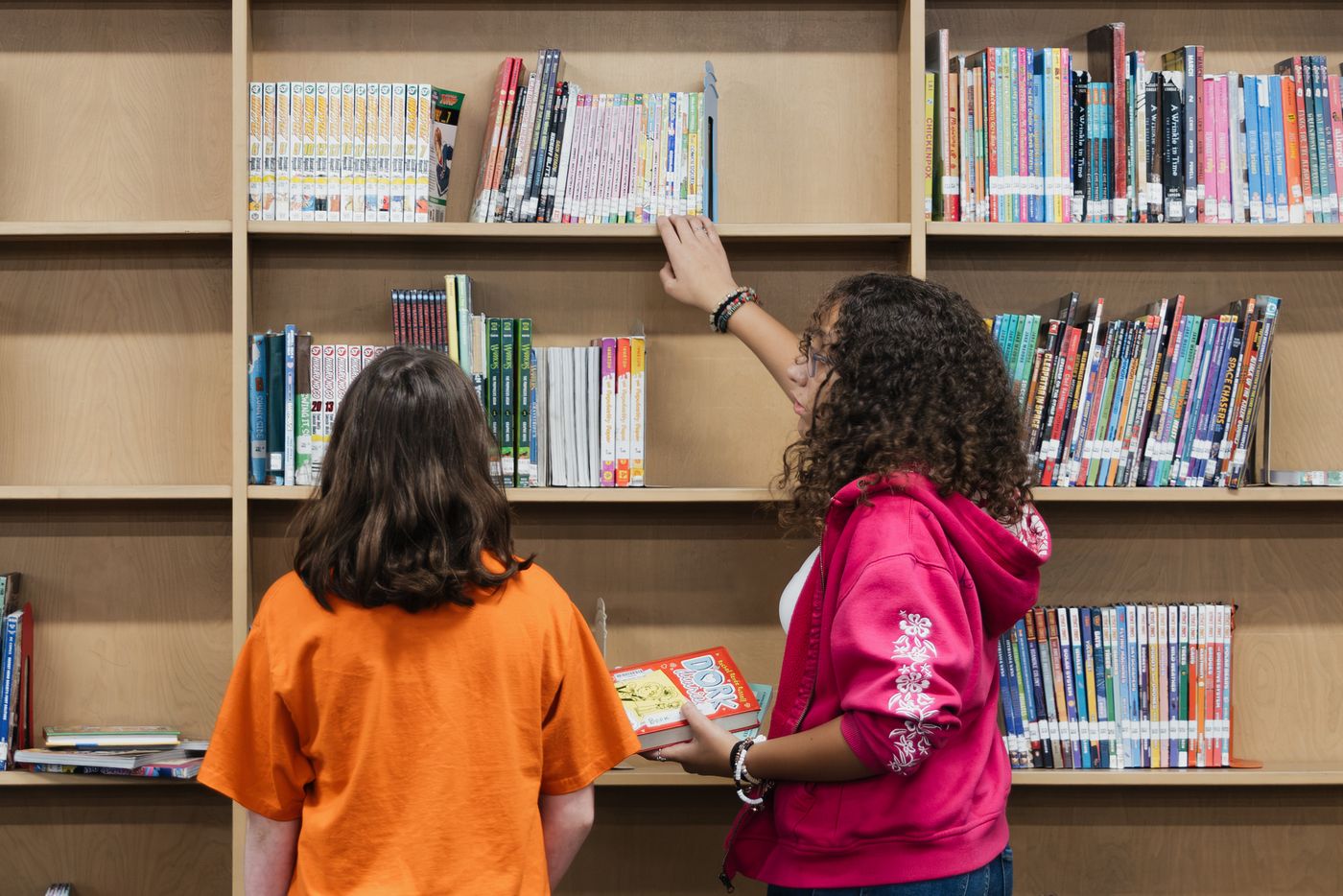 Two students reach for a book on a high library shelf.