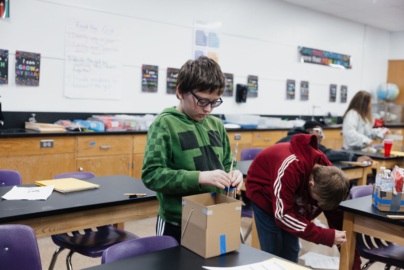 A student threads string into a cardboard box while a classmate works nearby.