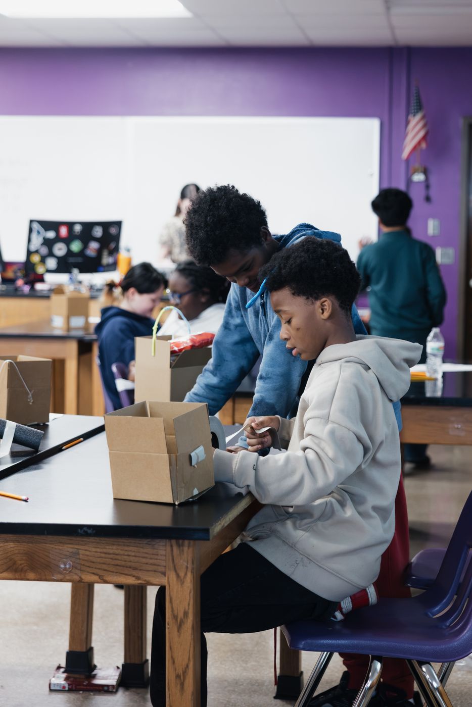 A student tapes a cardboard box together while a classmate helps.