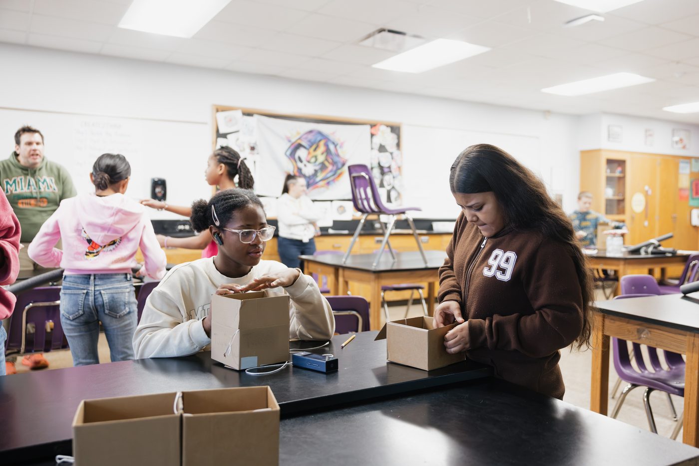 Two students fit out their cardboard boxes at a lab table.