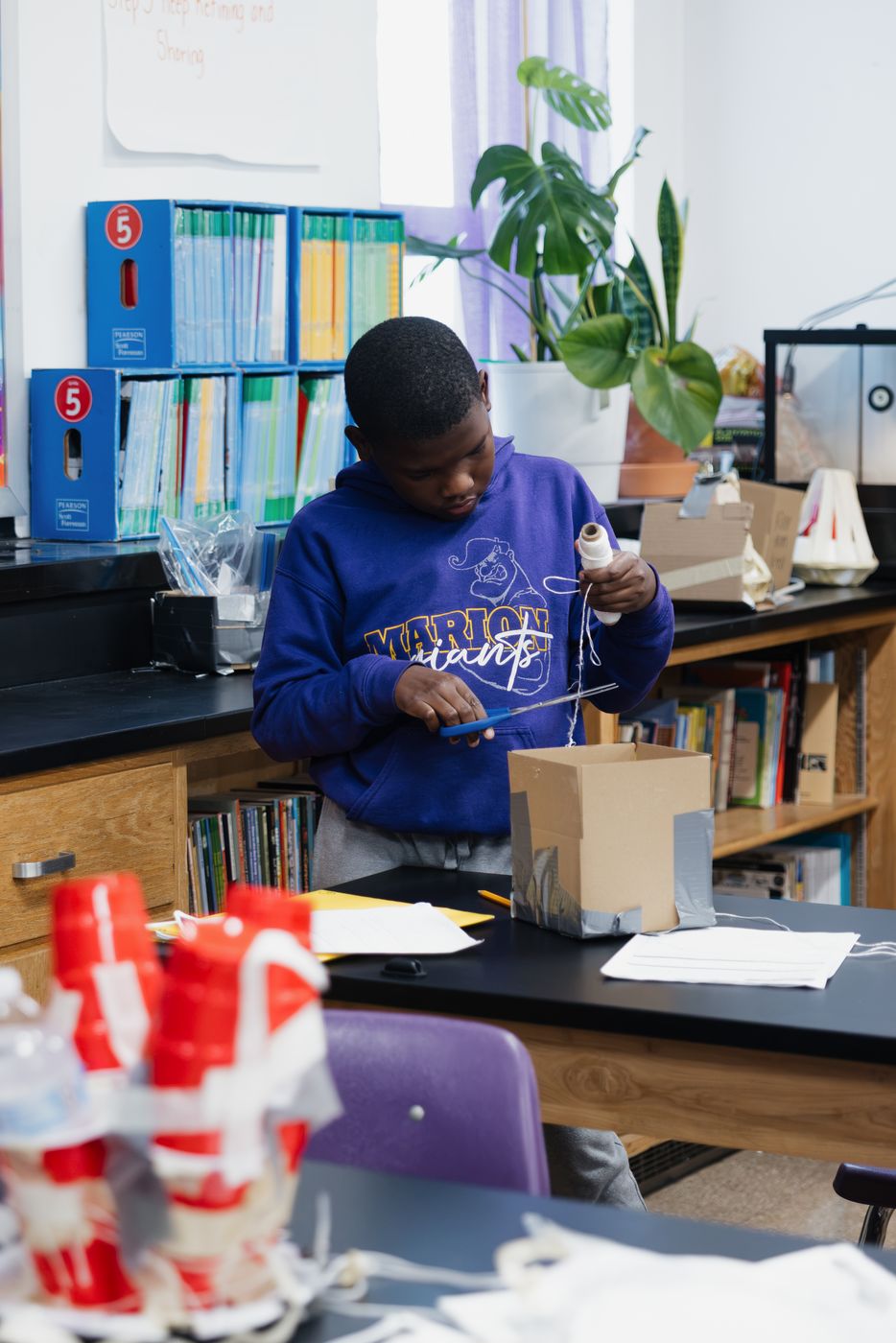 A student builds a cardboard device beside the classroom bookshelves.